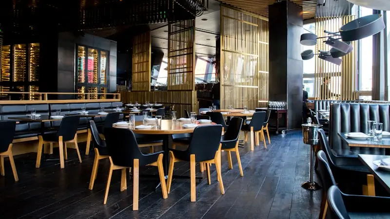 An empty modern restaurant dining room with navy chairs, pale wood tables and floor-to-ceiling brass screens.